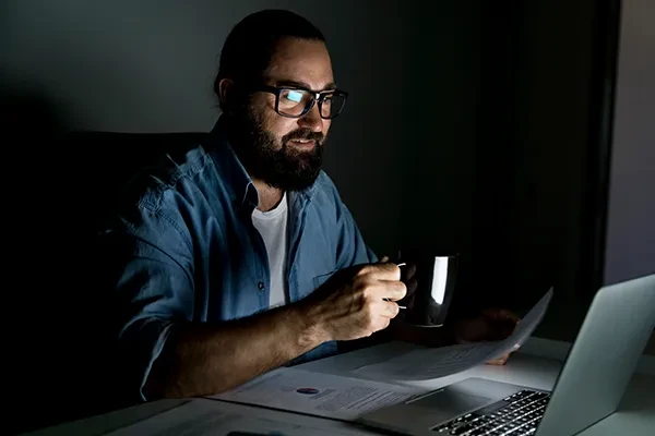Man with glasses holding mug in right hand and looking at laptop