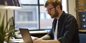 Man with glasses typing on laptop while sat at desk