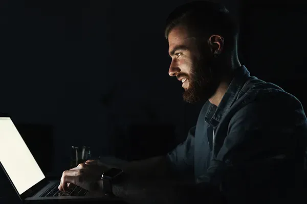 Man smiling while typing on laptop in dark