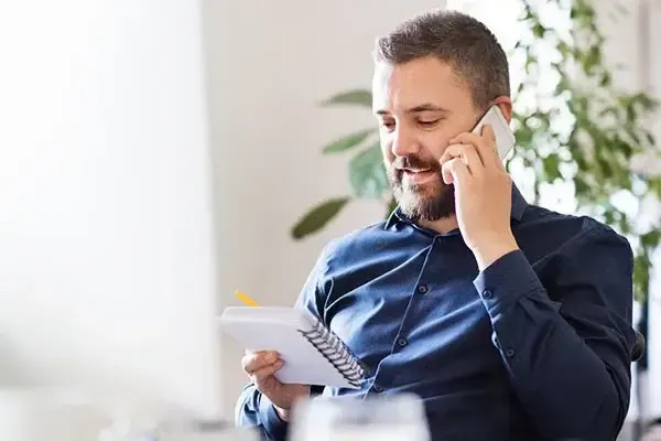 Man using a mobile phone in an office while reading a notepad