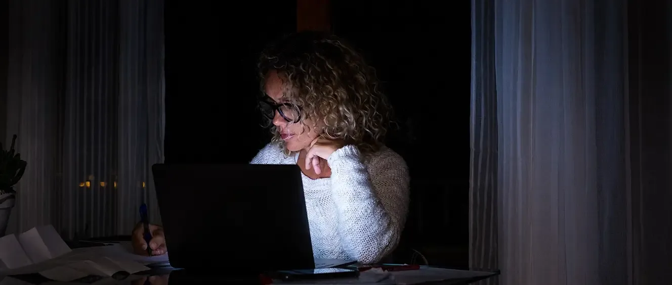 Woman with glasses looking at laptop while writing notes in dark room