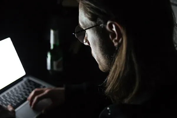 Man with glasses in dark room looking down at laptop