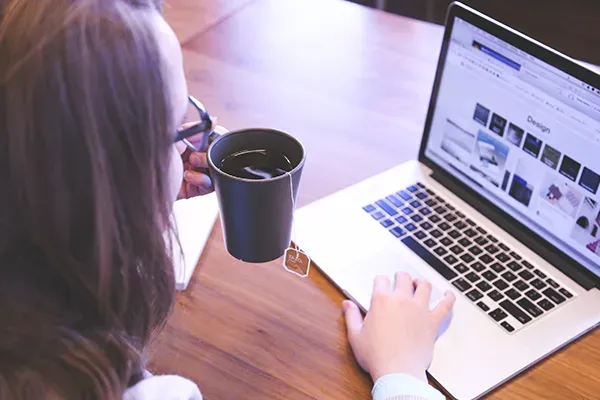 Woman with glasses holding coffee cup in left hand while typing on laptop